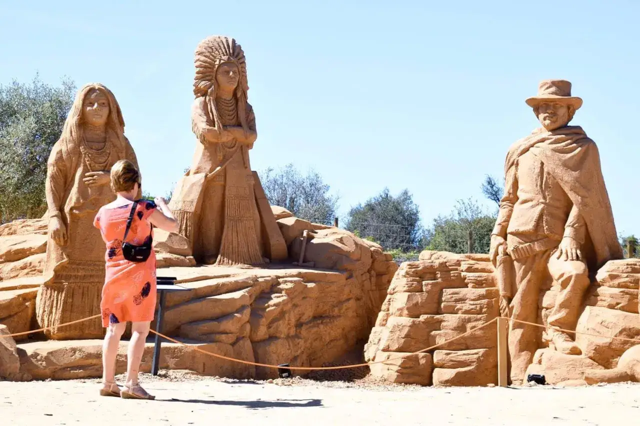 Sandskulpturen von Ureinwohnern und einem Cowboy in der Sand City Algarve. Eine Frau fotografiert die beeindruckenden Kunstwerke.