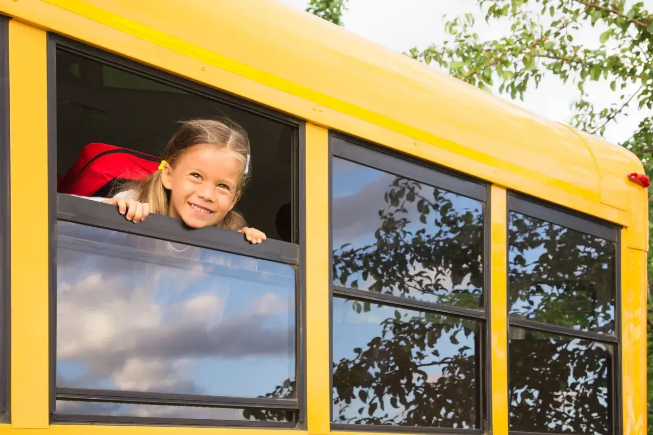 Une fillette souriante sort la tête par la fenêtre d'un bus scolaire jaune, prête pour son voyage. Une aide financière pour voyage scolaire rend cette aventure possible.