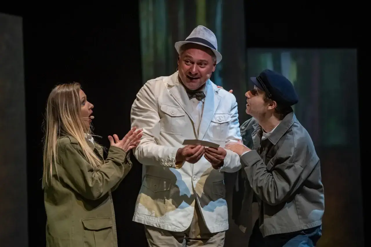 Trzech aktorów na scenie. Mężczyzna w białej marynarce i kapeluszu trzyma kartkę. Kobieta i mężczyzna w beretce patrzą na niego z zaciekawieniem. To streszczenie 