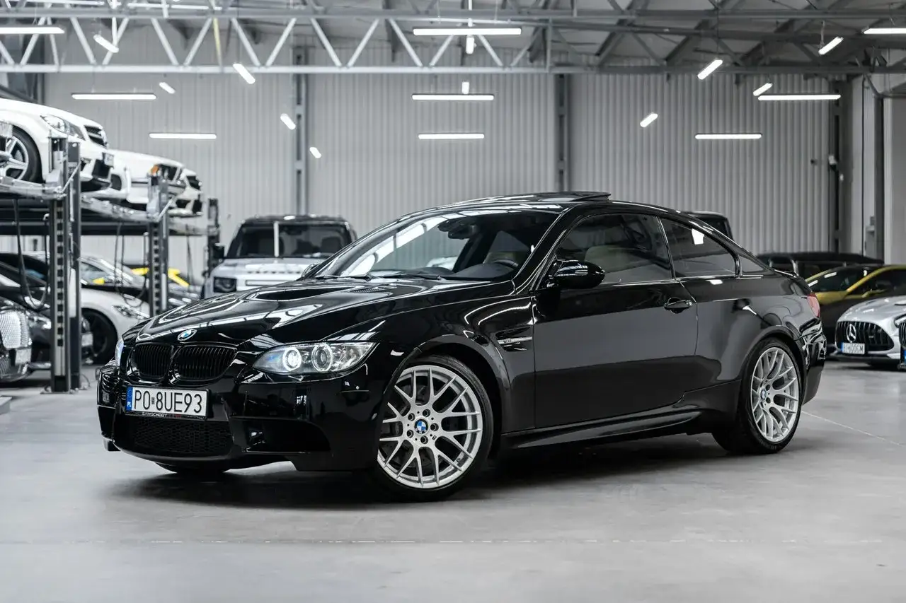 Błyszczące czarne BMW E90 M3 z felgami w stylu M. Samochód stoi w przestronnym garażu, obok innych luksusowych aut.