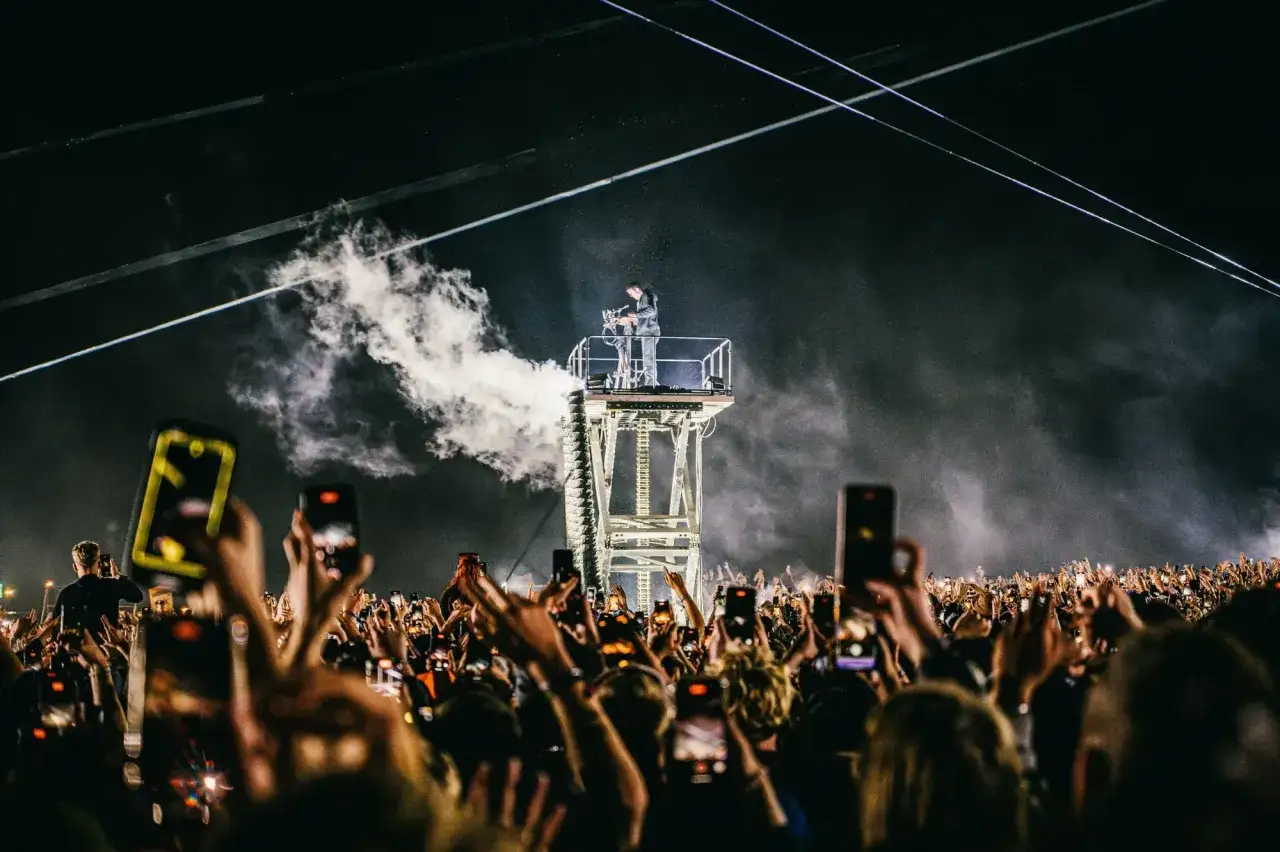 Un DJ pincha techno musica en una plataforma elevada, rodeado de humo y luces, mientras una multitud con móviles levanta las manos en señal de éxtasis.