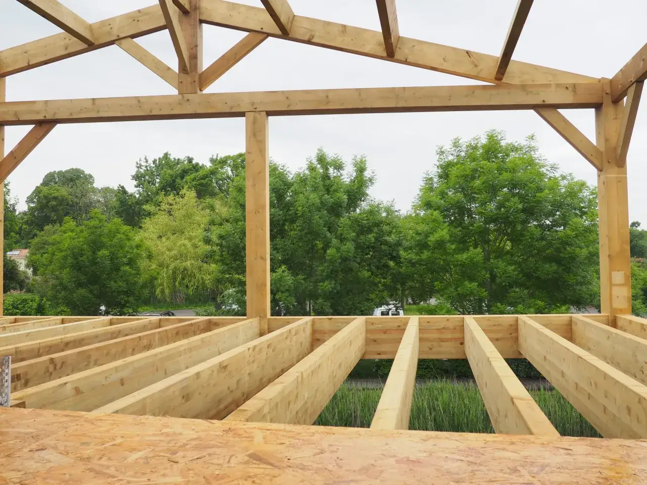 Vue d'une **structure en lamell&eacute; coll&eacute;** en cours de construction, avec des poutres en bois apparentes et une vue sur la verdure.