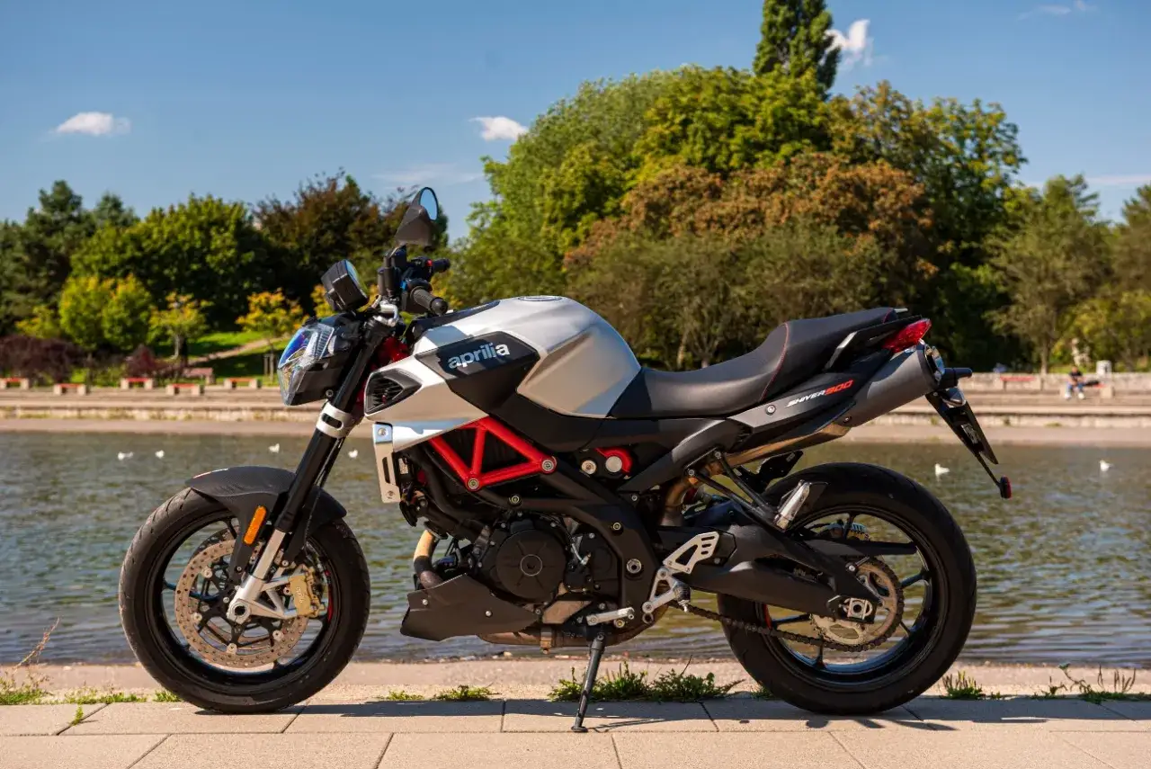 Srebrny motocykl Aprilia Shiver 900 z czerwonym stelażem stoi nad jeziorem w słoneczny dzień.