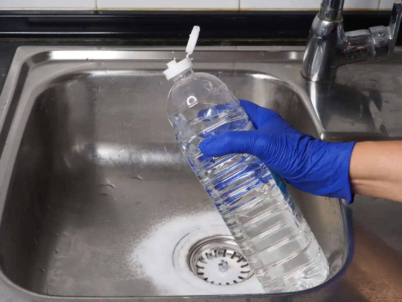Main tenant avec un gant bleu verse de l'eau d'une bouteille pour d&eacute;boucher l'&eacute;vier avec du percarbonate.