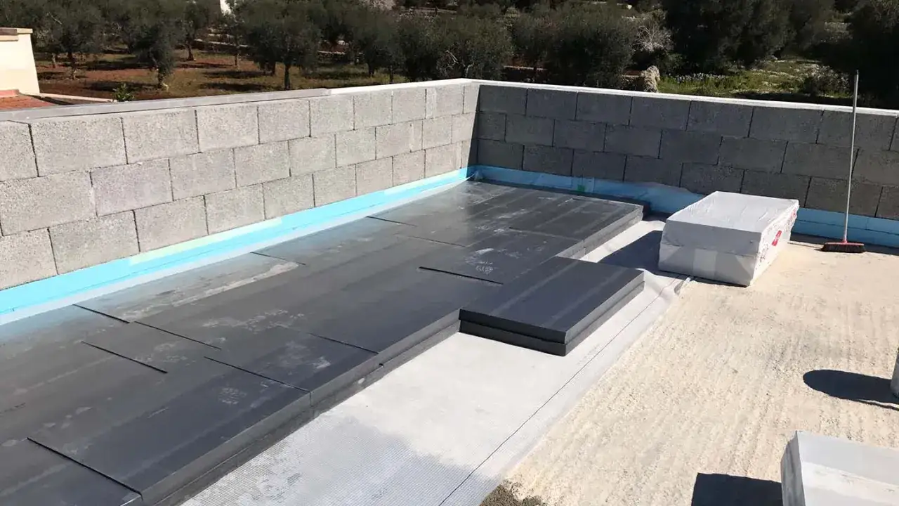 Posa di pannelli isolanti e materiali per impermeabilizzazioni edili su un tetto in costruzione, con muretto in blocchi di cemento.