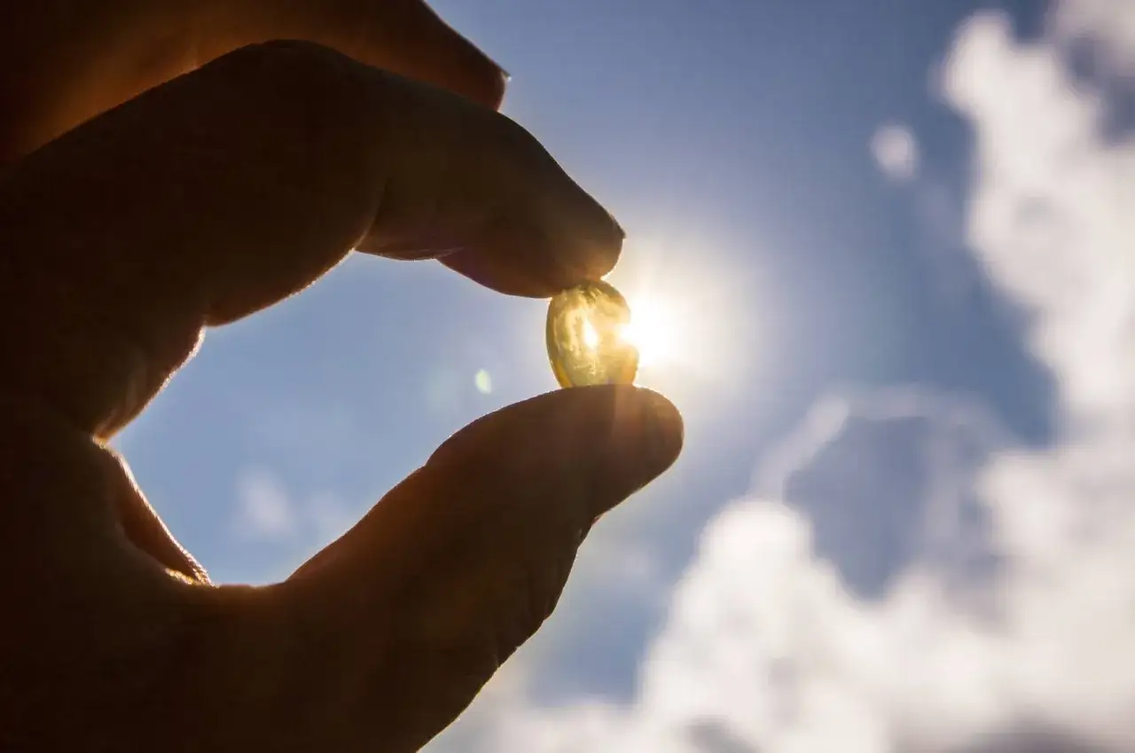 En hand håller upp en solbelyst kapsel mot en blå himmel med moln. Kapseln symboliserar vitamin D och dess viktiga funktion för kroppen.