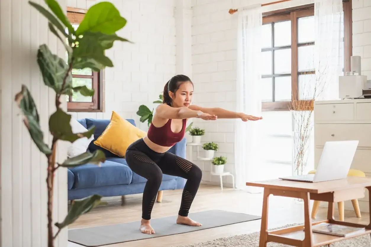 Donna esegue squat a casa, seguendo un tutorial online. Il suo allenamento mira a tonificare il corpo, migliorando forza e benessere.