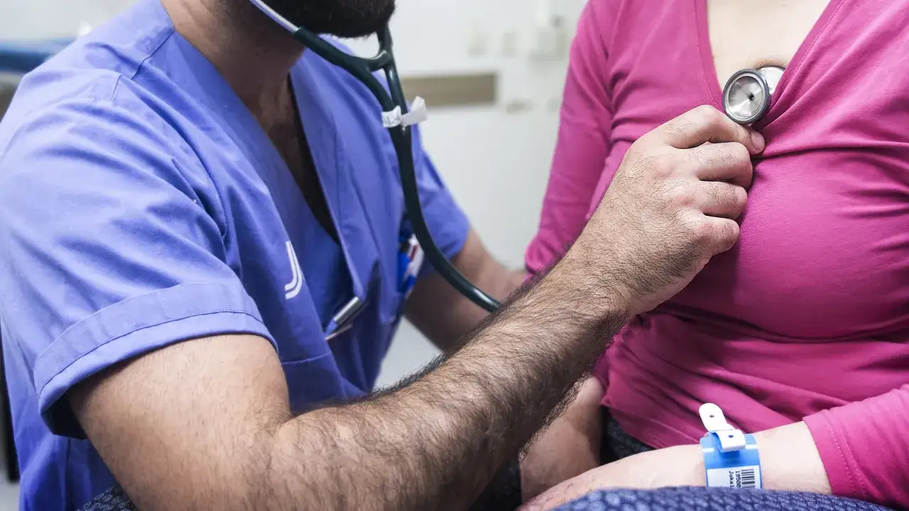 L&auml;kare lyssnar p&aring; hj&auml;rtat med stetoskop. Patienten har ett armband, kanske f&ouml;r att f&aring; sjukskrivning vid utmattningssyndrom.