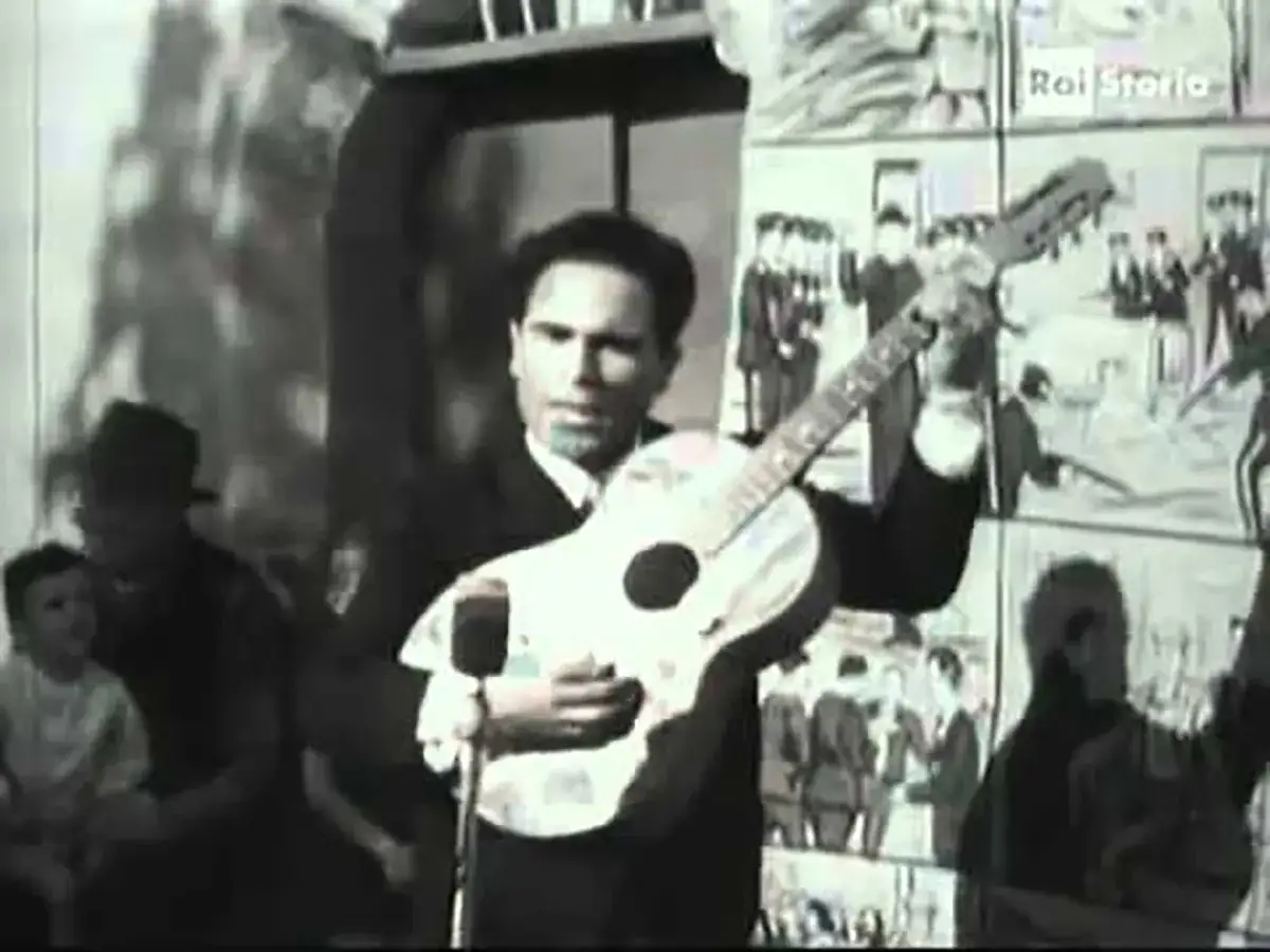 Uomo con chitarra, forse canta una canzone in dialetto siciliano, con pubblico sullo sfondo.