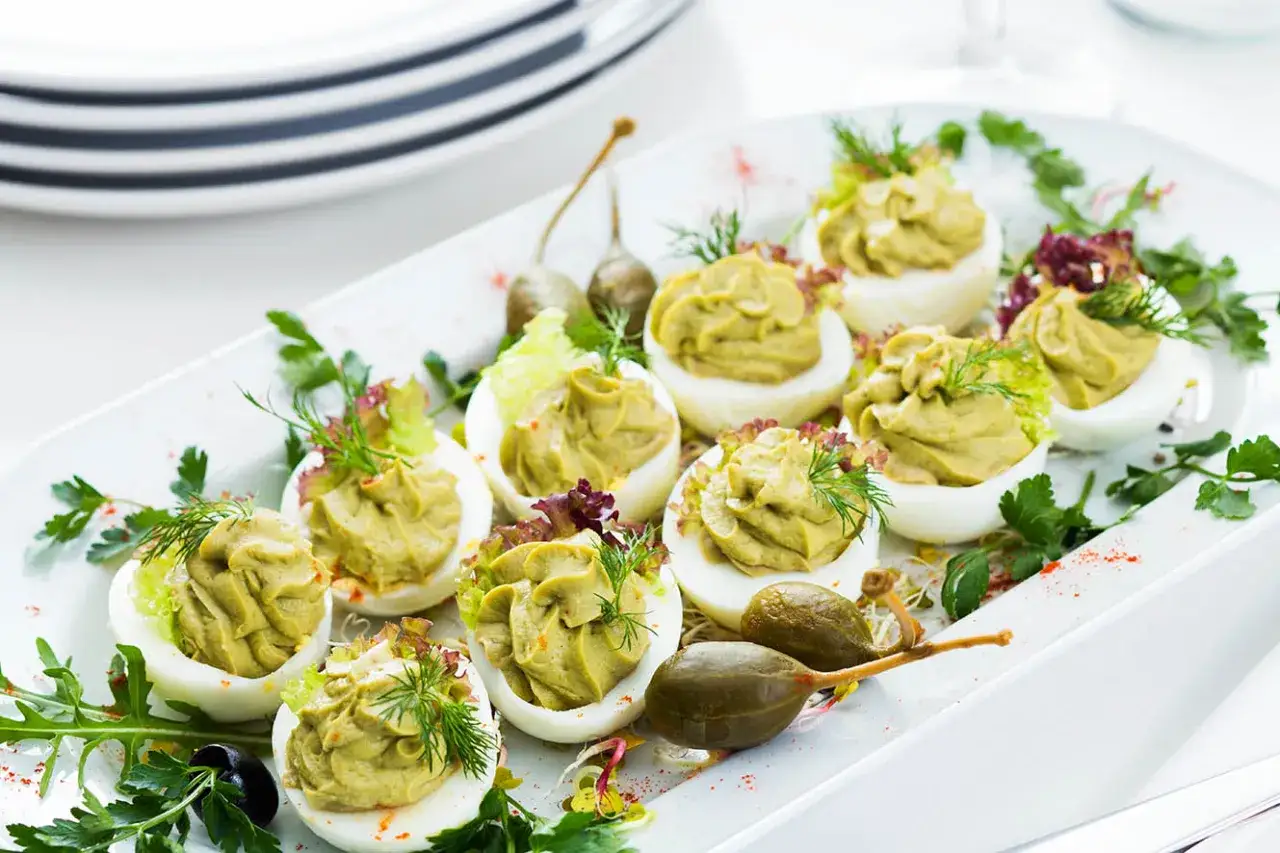 Jajka faszerowane guacamole, udekorowane zieleniną i kaparami, prezentują się apetycznie na białej tacy.