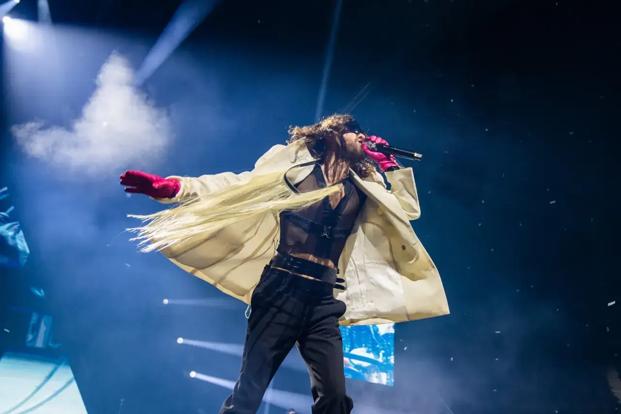 Jared Leto z zespołem na scenie, w białym płaszczu i różowych rękawiczkach, śpiewa do mikrofonu.