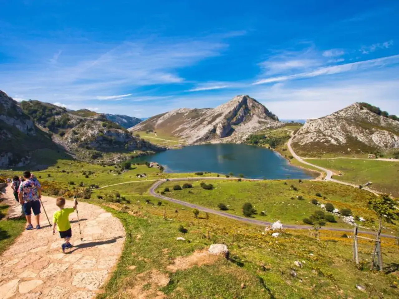 Un niño con bastones de senderismo camina por un sendero de piedra hacia un lago azul rodeado de montañas. ¡Descubre qué ver en Picos de Europa!