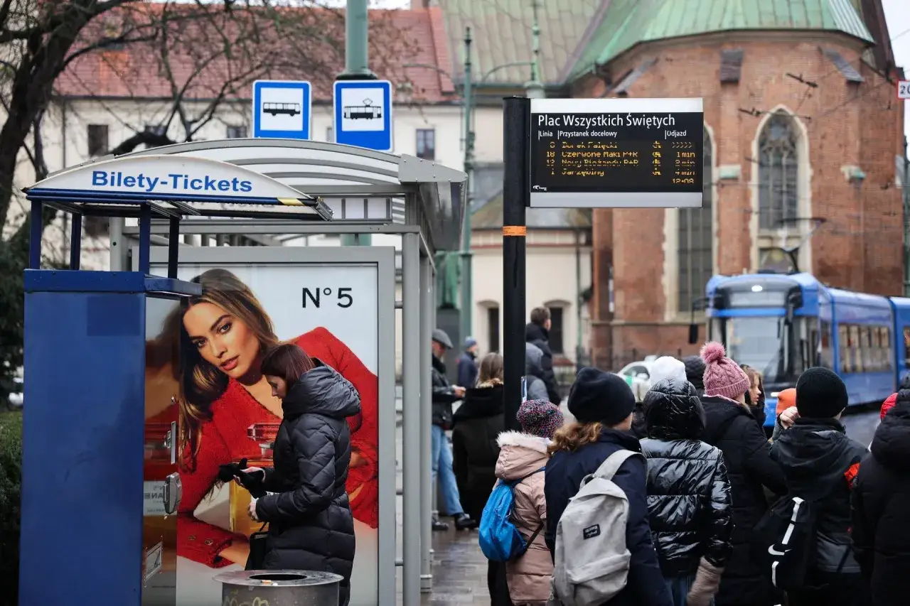 Przystanek tramwajowy z tablicą odjazdów i reklamą. Ktoś kupuje bilet miesięczny, by pojechać tramwajem.