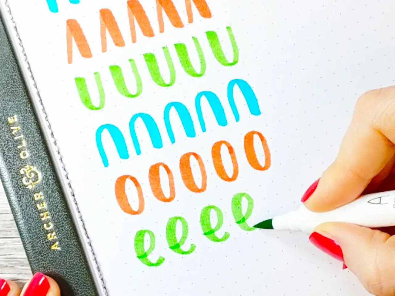 Manos con u&ntilde;as rojas practicando lettering con rotulador verde en cuaderno de puntos.