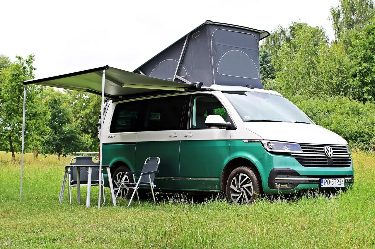 Zielony kamper VW California z rozłożonym namiotem dachowym i markizą, obok stolik i krzesła. Idealny na wakacje, poznaj jego vw california wymiary.