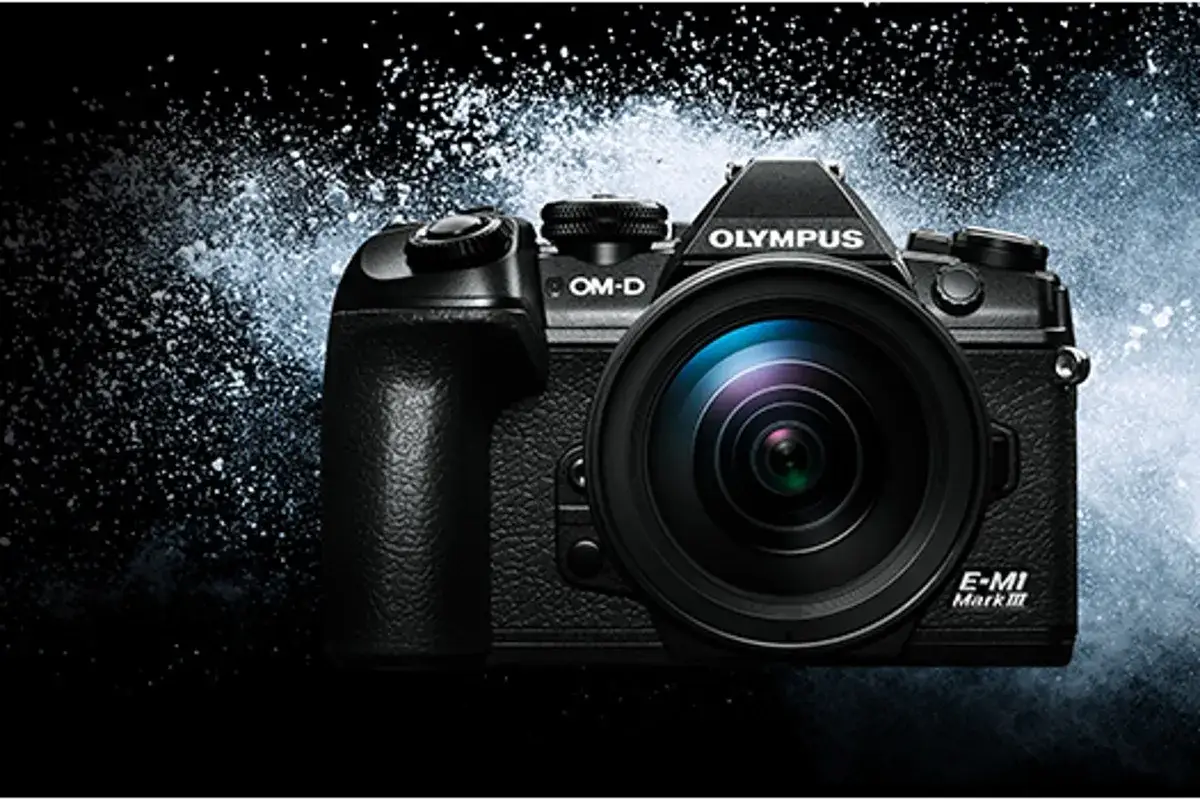 Olympus E-M1 Mark III, una fotocamera mirrorless con obiettivo, circondata da una nuvola di polvere bianca su sfondo scuro.