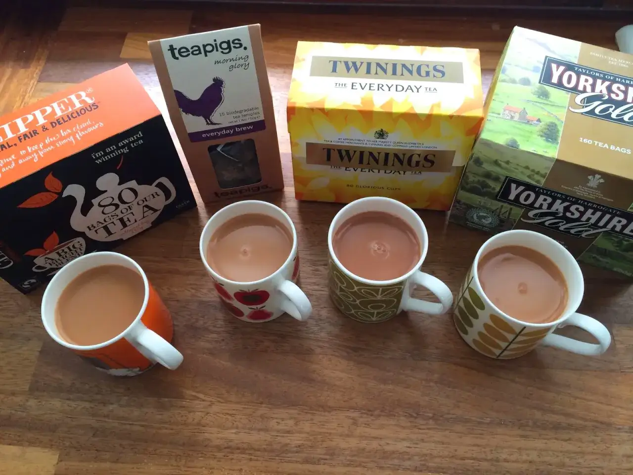 Ekskluzywne herbaty w saszetkach: Teapigs, Twinings i Yorkshire Gold. Cztery kubki z parującą herbatą na drewnianym stole.