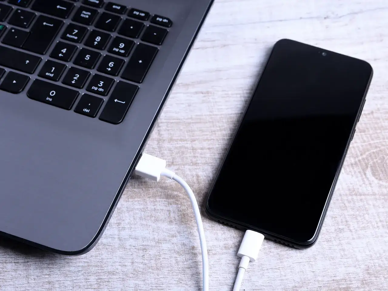 Jak połączyć telefon z laptopem? Widok z góry na podłączony kablem biały kabel USB do czarnego smartfona i szarego laptopa.