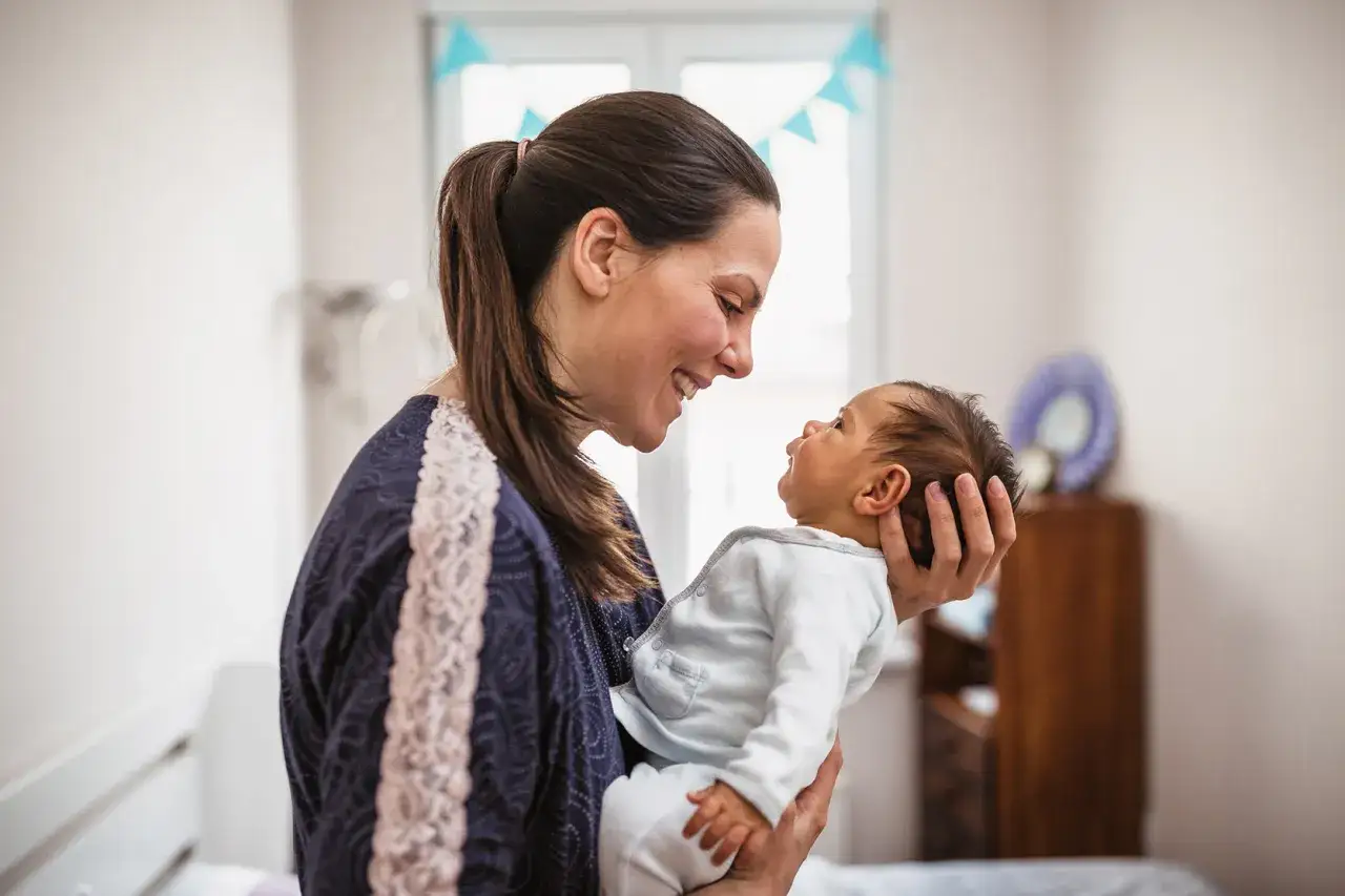 Uśmiechnięta mama tuli noworodka. Dziecko patrzy na nią z zaciekawieniem, jakby miało zaraz kichnąć.