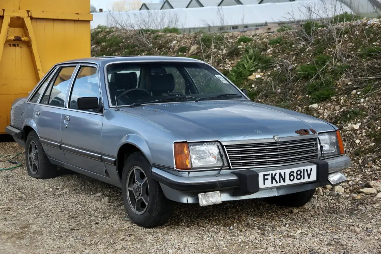 En ljusbl&aring; Vauxhall Firenza st&aring;r parkerad p&aring; grus. Bilen har en rostfl&auml;ck p&aring; huven och en punktering.