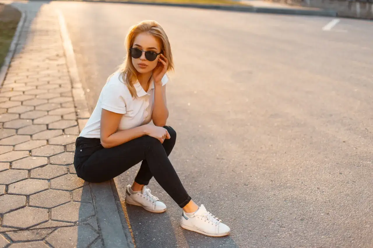 Blondynka w czarnych jeansach i białej koszulce polo, siedząca na chodniku. Stylowa inspiracja, jak nosić koszulka polo damska na co dzień.