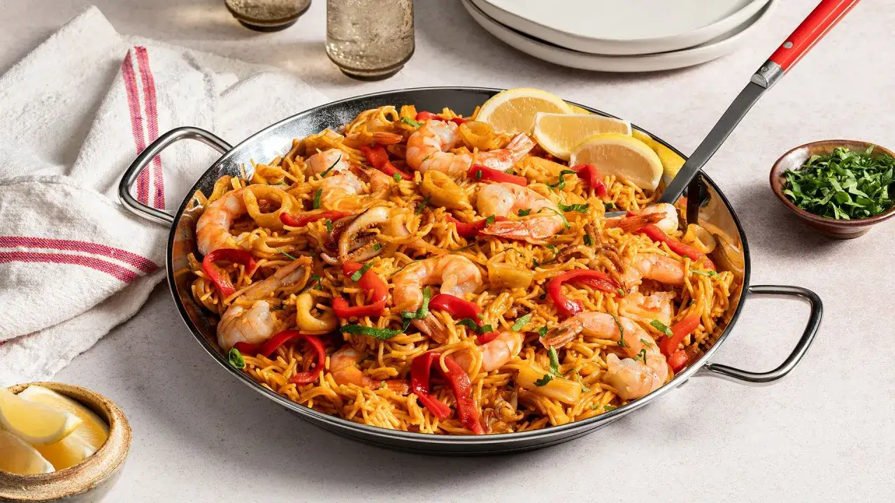 Fideos fideuá con gambas, calamares y pimientos rojos, servido en paellera con gajos de limón y perejil picado.