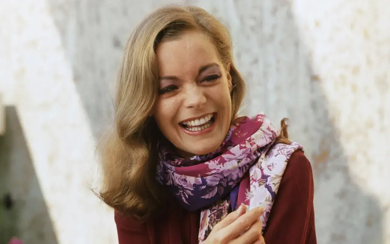 Uśmiechnięta kobieta z blond włosami i kolorowym szalem, przypominająca francuskie gwiazdy porno z lat 70.