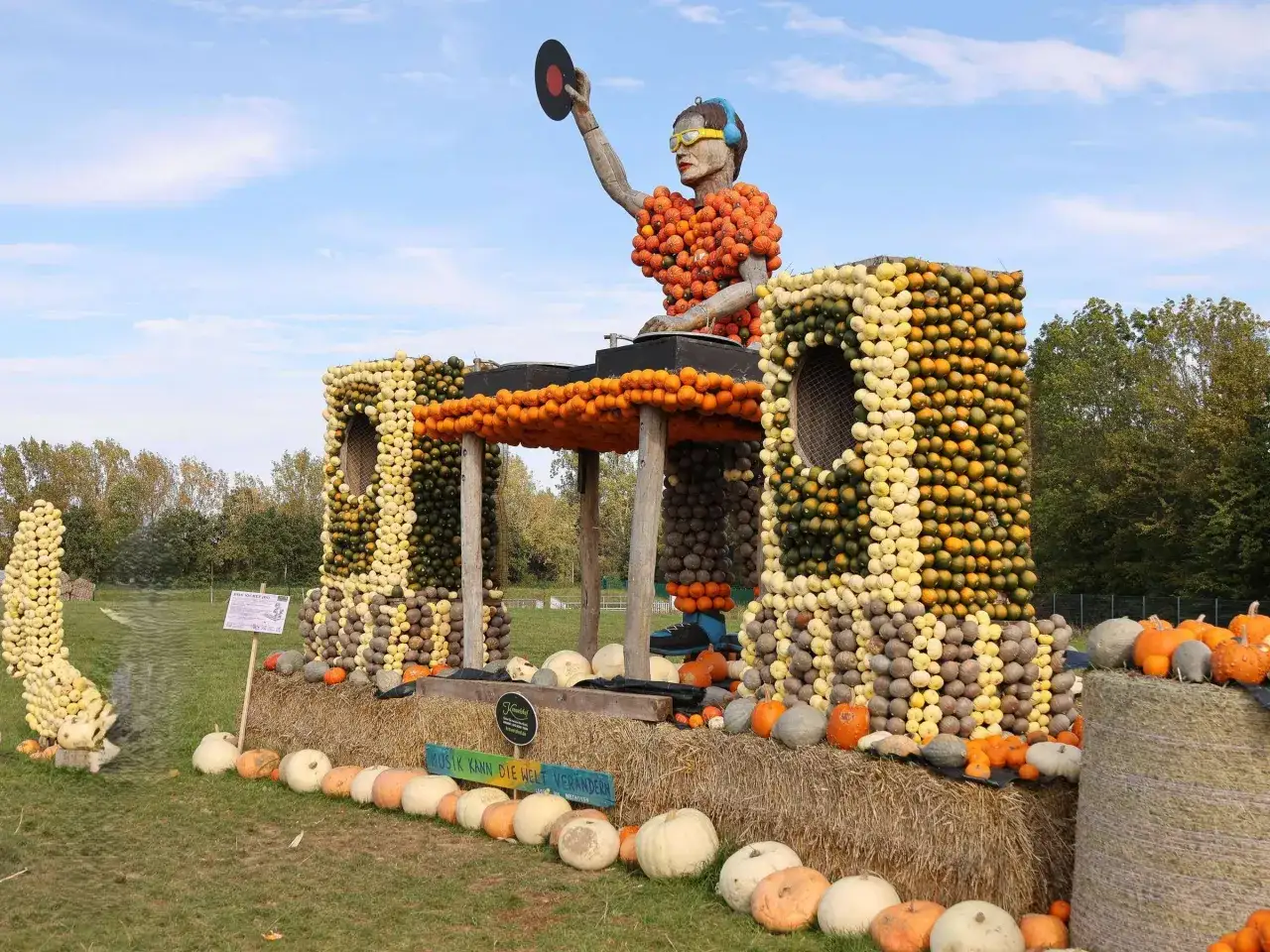 DJ aus Kürbissen auf einem herbstmarkt nrw, der eine Schallplatte hält. Bunte Kürbis-Skulpturen und Heuballen schaffen eine festliche Atmosphäre.