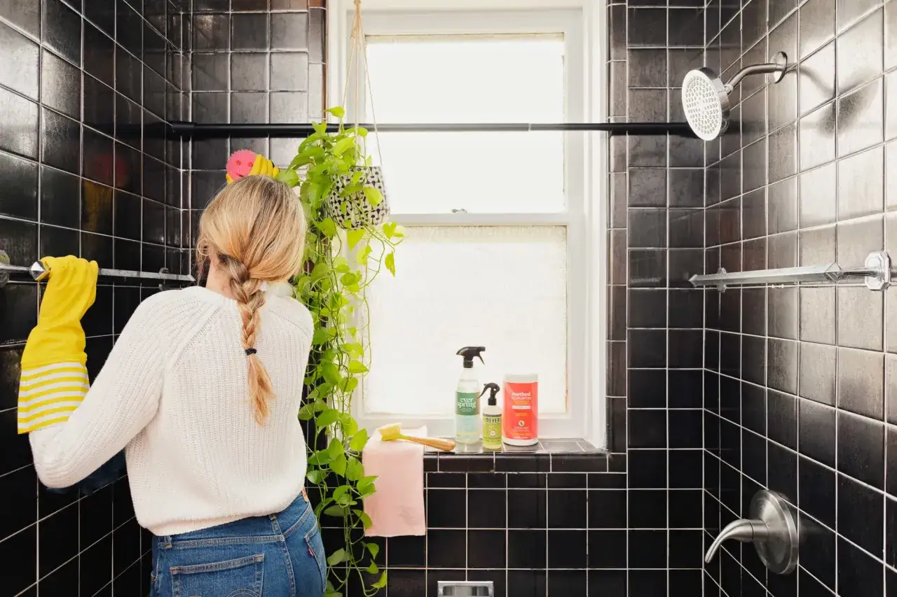 Woman in yellow gloves scrubs black tiled shower, tackling bathroom mold with cleaning supplies.