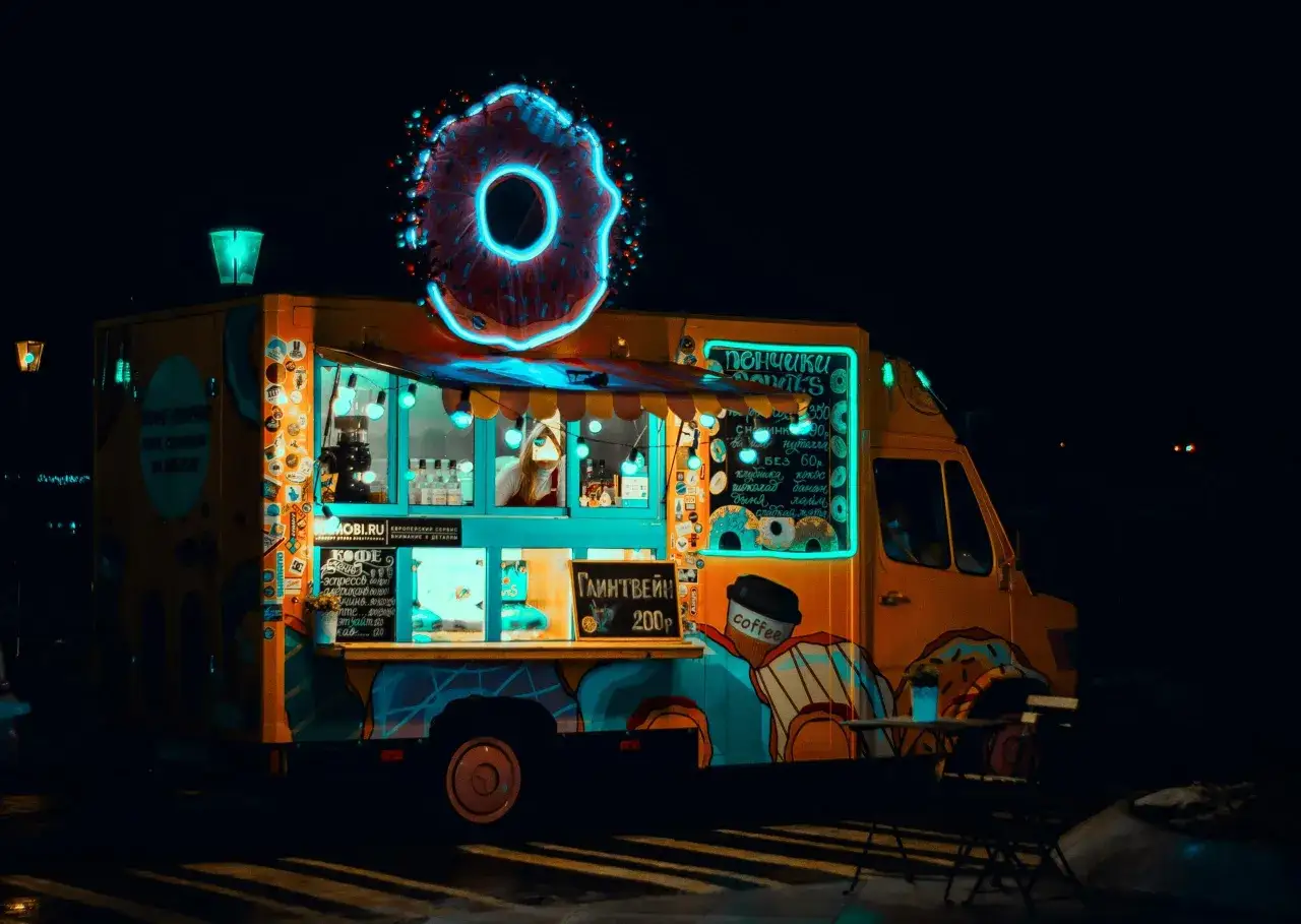 Nocny food truck z pączkami i kawą, ozdobiony neonowym pączkiem. Menu wskazuje, że glintwein kosztuje 200p.