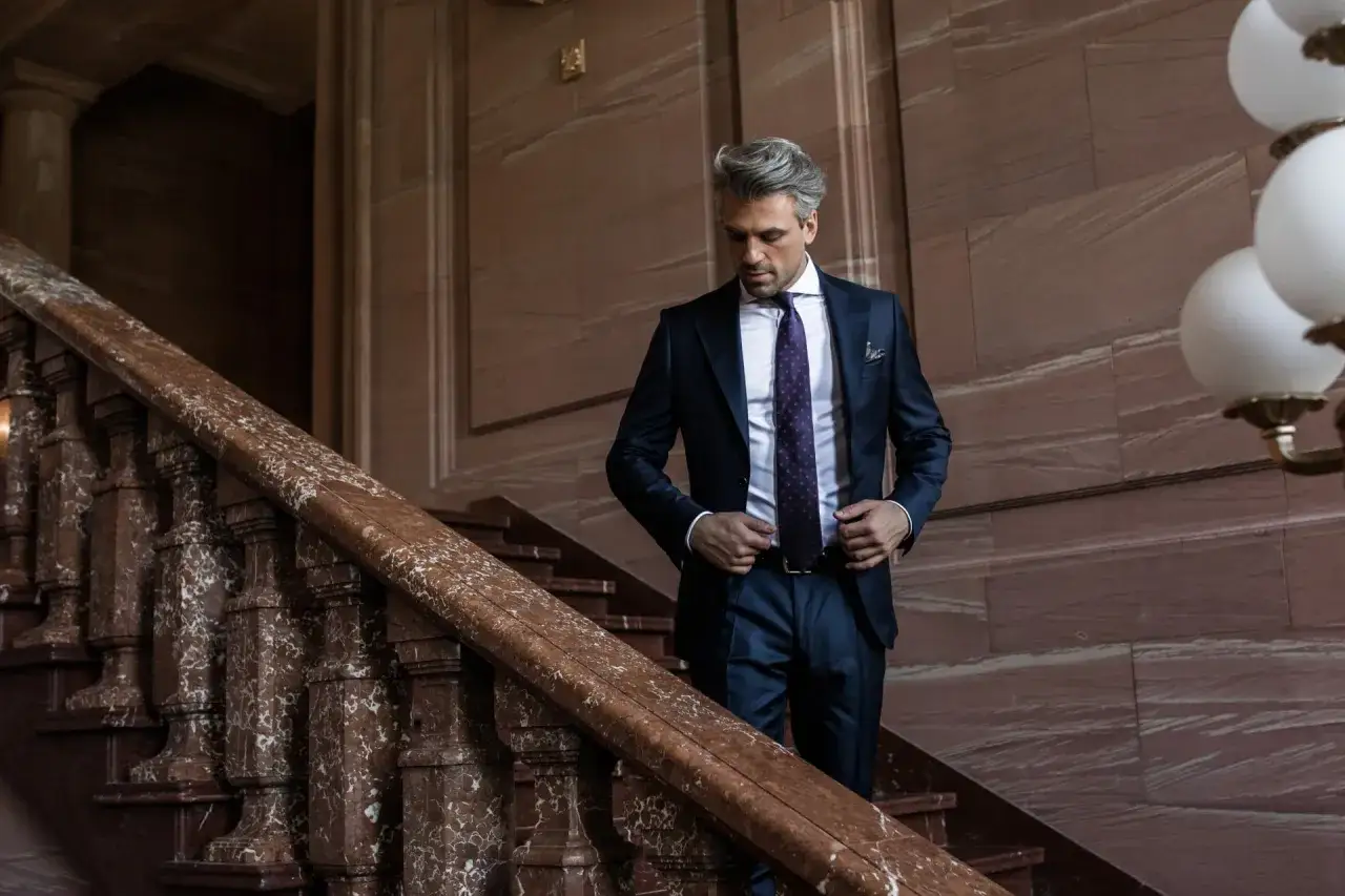Elegancki mężczyzna w granatowym garniturze i fioletowym krawacie schodzi po marmurowych schodach. Idealne pytanie: jakie skarpety do granatowego garnituru?