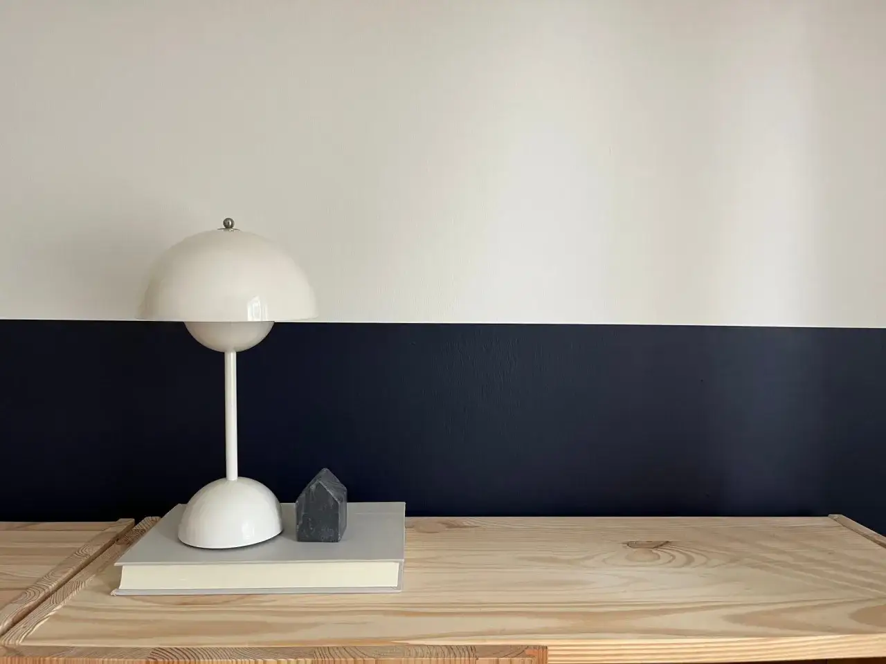 Lampe blanche sur un livre, devant un mur deux couleurs différentes : blanc et bleu nuit. Un petit objet gris en forme de maison est posé sur le livre.