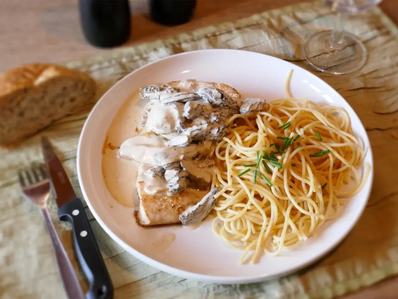 Un plat de pâtes spaghetti avec du poulet nappé d'une onctueuse sauce aux morilles. Un morceau de pain et des couverts sont à côté.