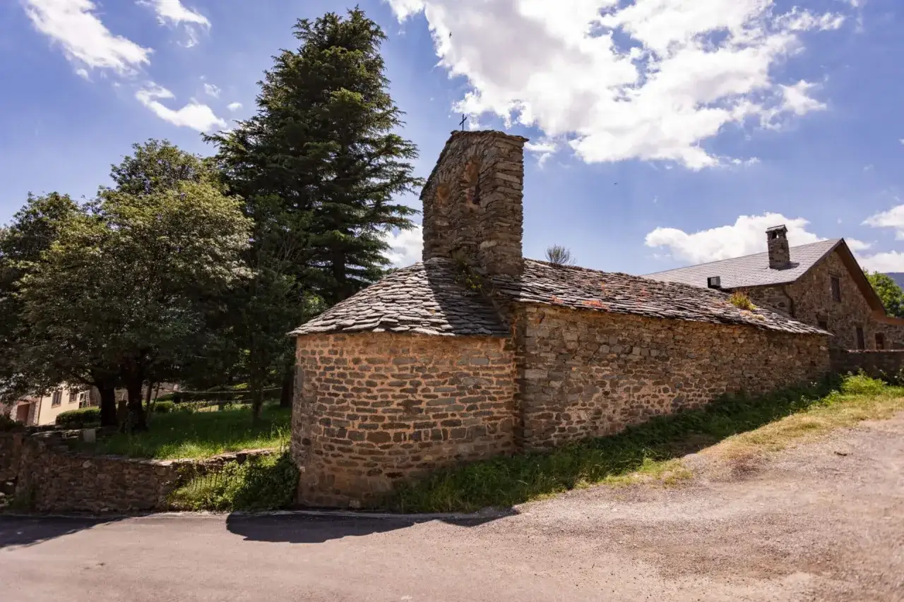 Iglesia de piedra con tejado de pizarra en el Ripollès, rodeada de vegetación y bajo un cielo azul con nubes.