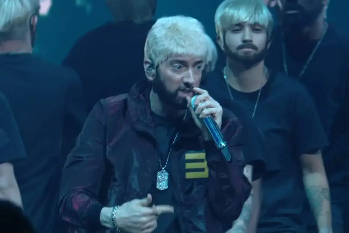 Slim Shady na scenie, w błyszczącej kurtce, z mikrofonem w dłoni. Otoczony przez ludzi z blond włosami.