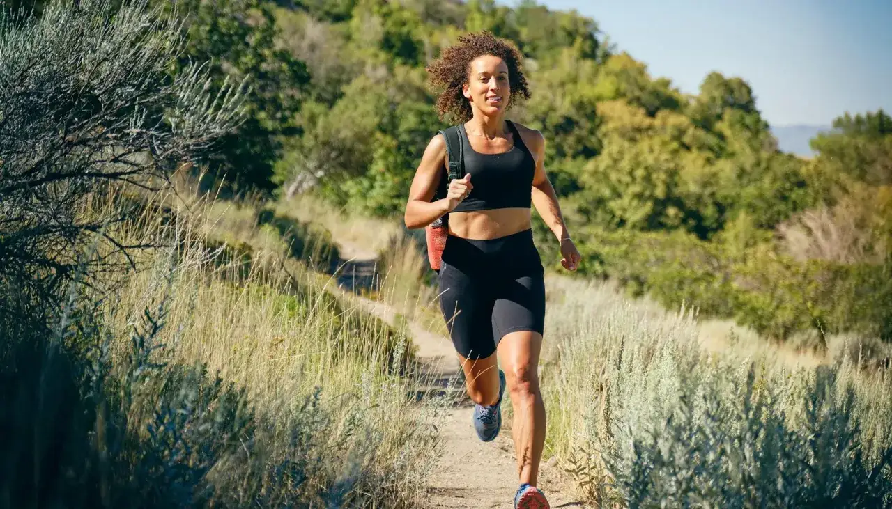 Frau joggt auf einem Feldweg durch h&uuml;gelige, gr&uuml;ne Landschaft. Sie tr&auml;gt Sportkleidung und einen Rucksack. Joggen ist gesund!
