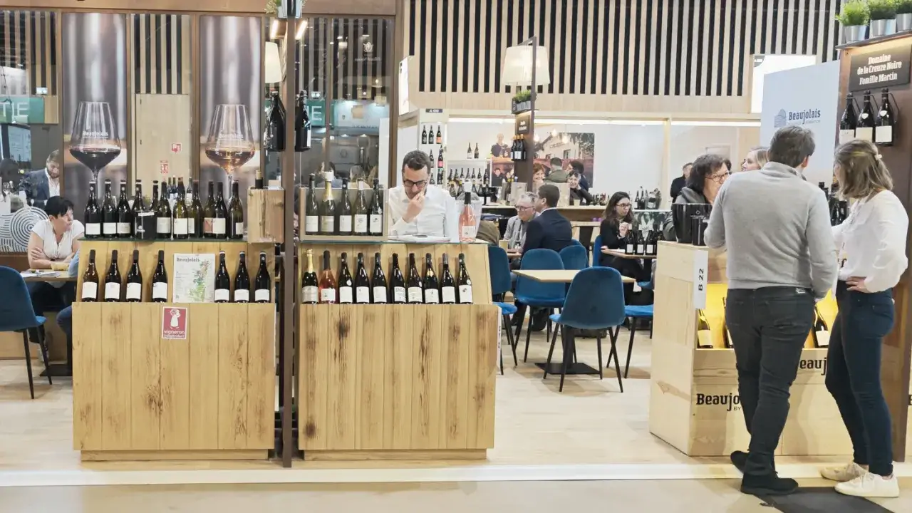 Salon du vin à Paris en 2026 : dégustation de bouteilles sur un stand en bois, avec des visiteurs discutant et des tables bleues.