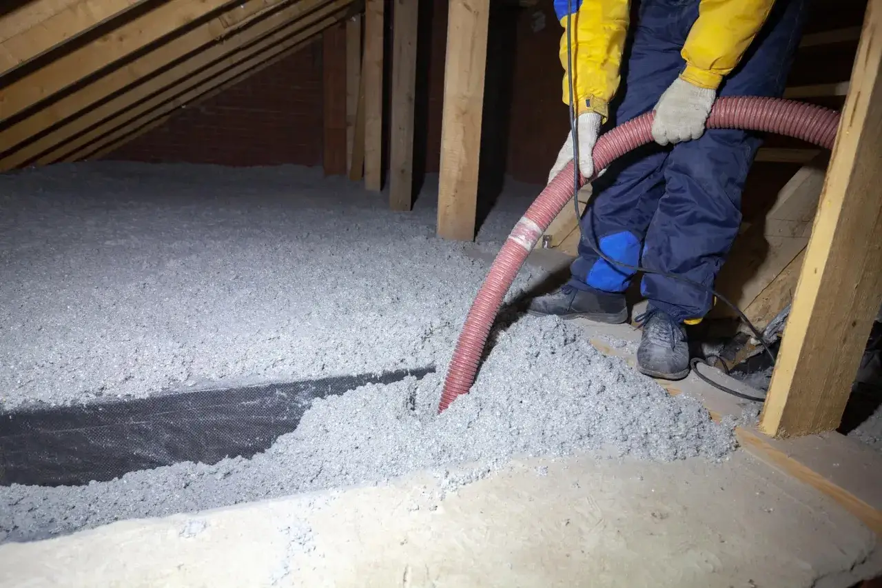 Un ouvrier installe de l'isolant en **cellulose** dans un grenier, soufflant le matériau par un tuyau rouge.
