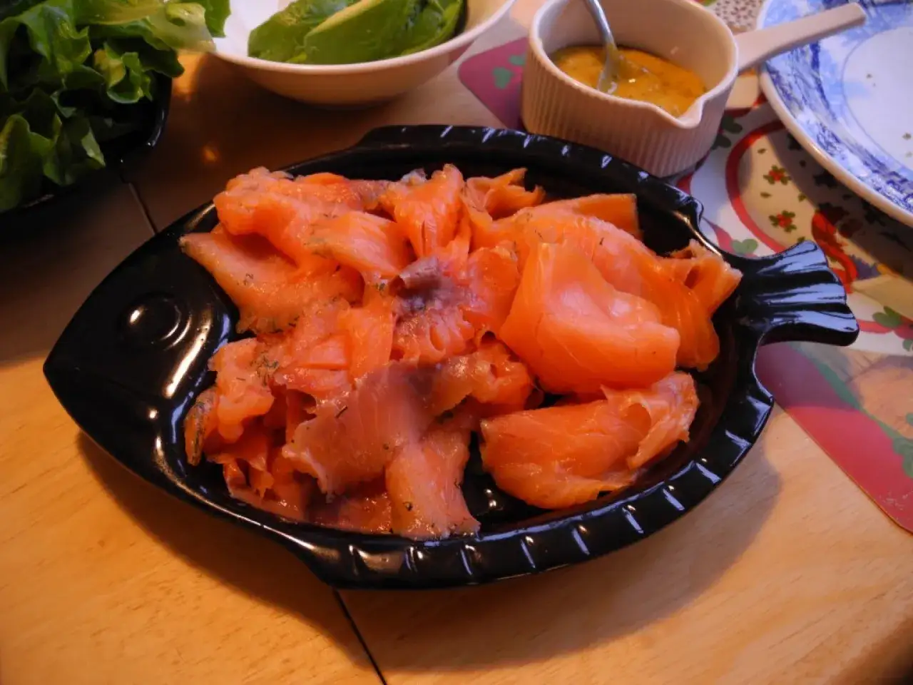 En svart fiskformad skål fylld med läcker rimmad lax, strödd med dill. Bredvid finns en liten skål med senapssås och en skål med sallad.