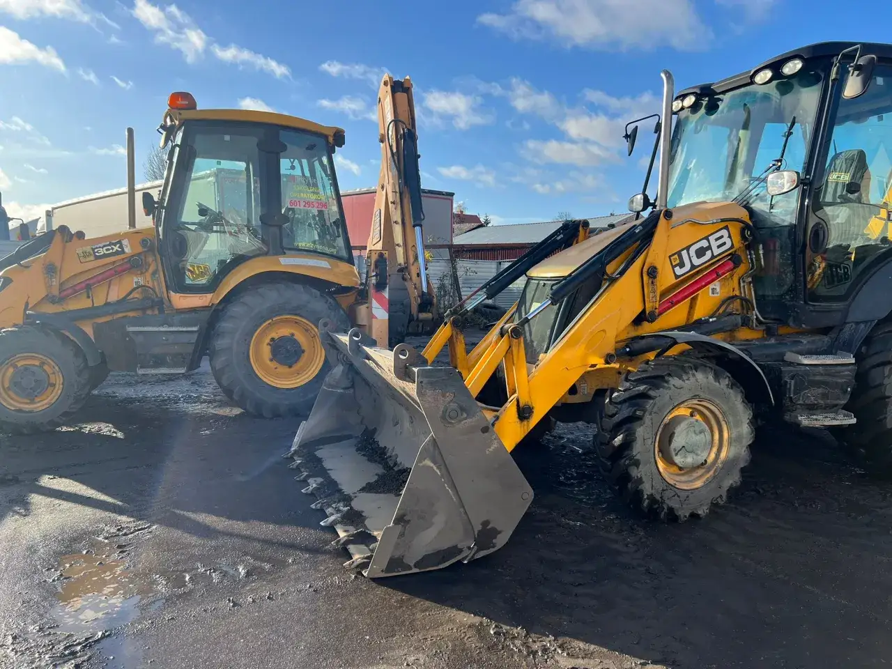 Dwie żółte koparki JCB stoją na błotnistym placu. Jedna z nich ma na szybie napis