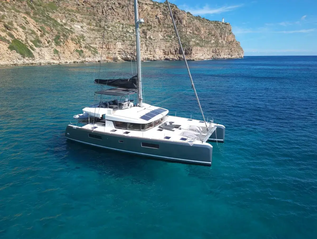 Ce catamaran moderne, idéal pour traverser l'Atlantique, flotte dans des eaux turquoise cristallines, avec une falaise rocheuse et un phare en arrière-plan.