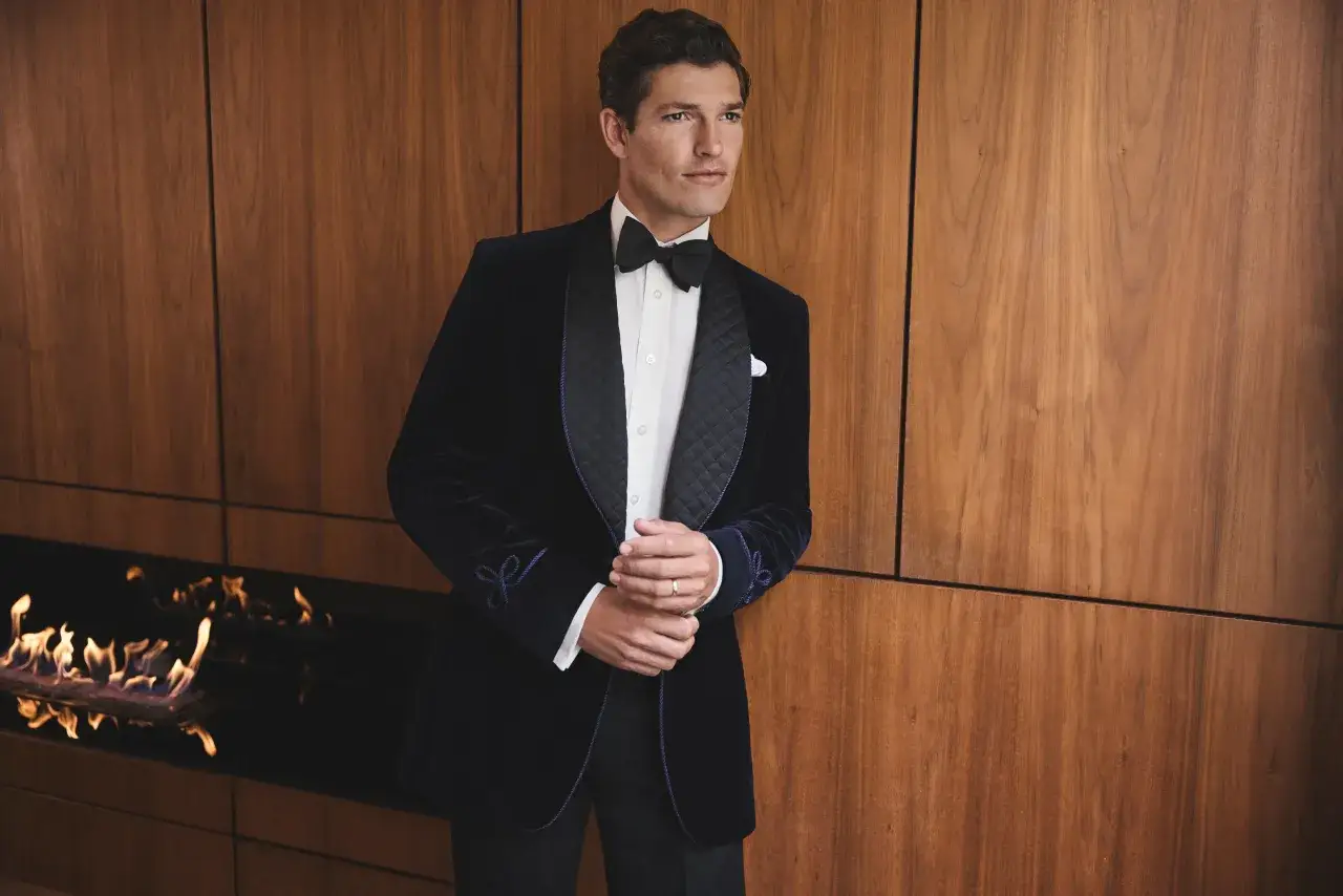 A man in a festive dress code, sporting a velvet tuxedo with a bow tie, stands by a modern fireplace.