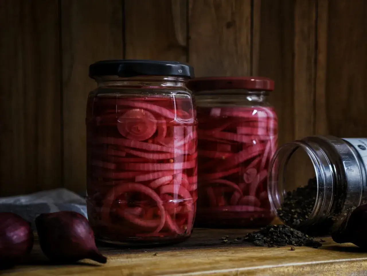 Zwei Gläser mit eingelegten roten Zwiebeln stehen auf einem Holztisch. Daneben liegen frische rote Zwiebeln und ein Glas mit Pfefferkörnern.