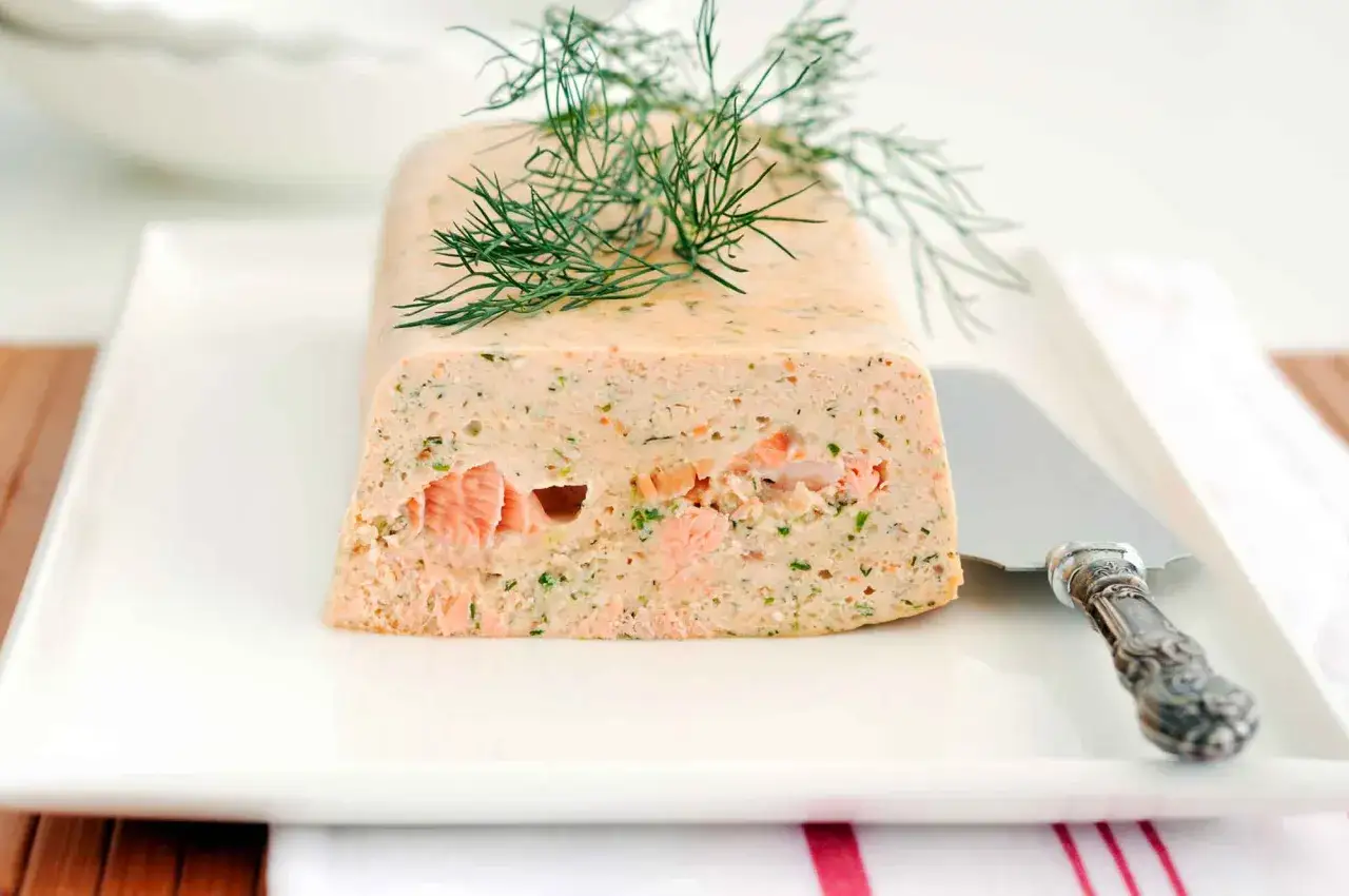 Une tranche de terrine de poisson, garnie d'aneth frais, sur une assiette blanche avec un couteau à découper.