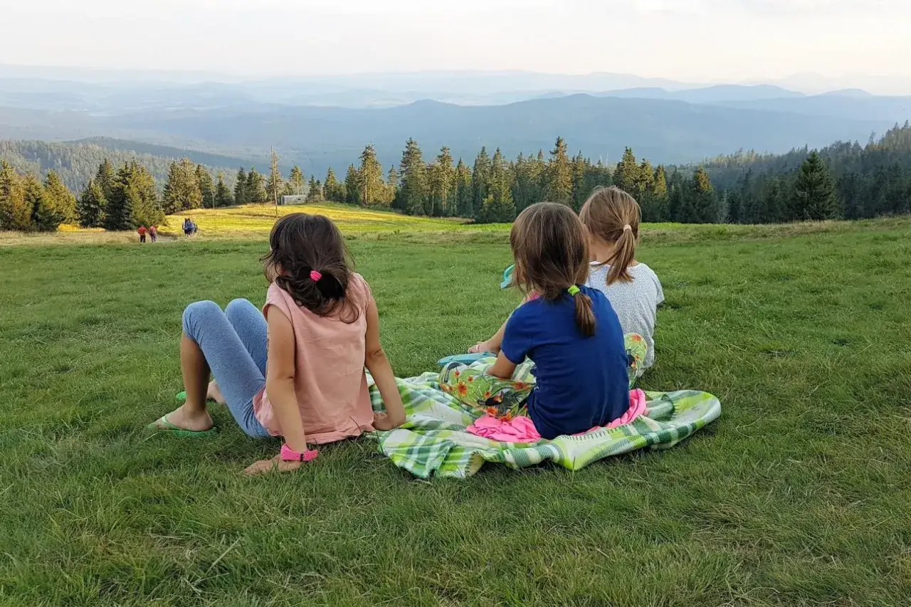 Trzy dziewczynki odpoczywają na trawie, podziwiając widok gór. To idealne miejsce na odpoczynek po wędrówce rysianka szlaki z dzieckiem.