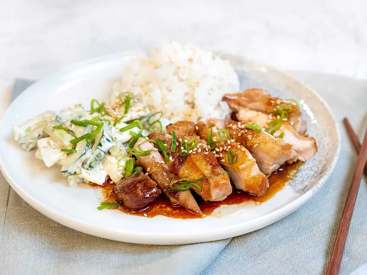 En tallrik med saftig teriyaki kyckling, ris och en krämig gurksallad. Strössel av sesamfrön och grön lök ger extra smak.