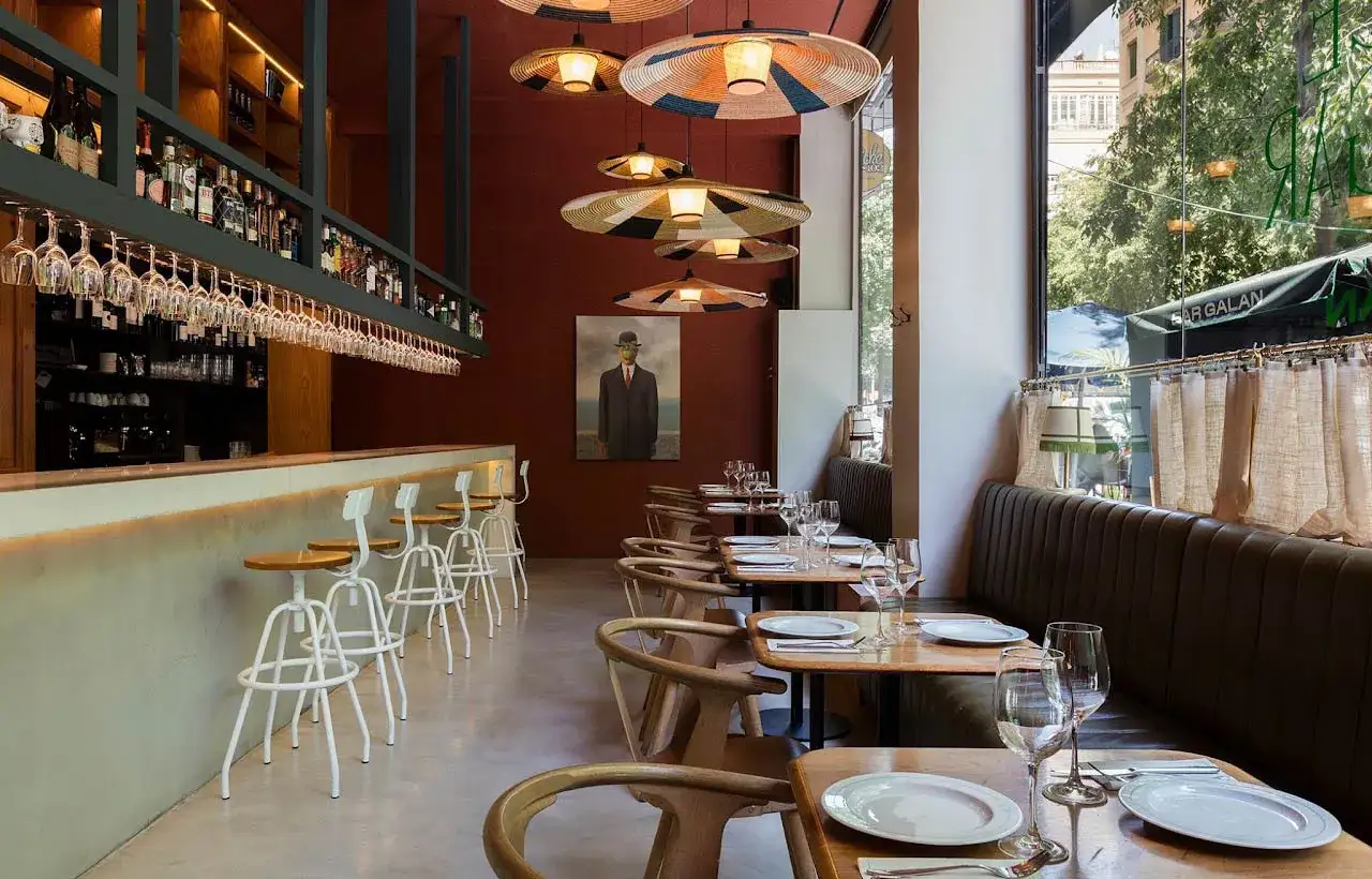 Elegante decoracion restaurante con barra, mesas de madera y cuadros. Lámparas colgantes y asientos cómodos crean un ambiente acogedor.
