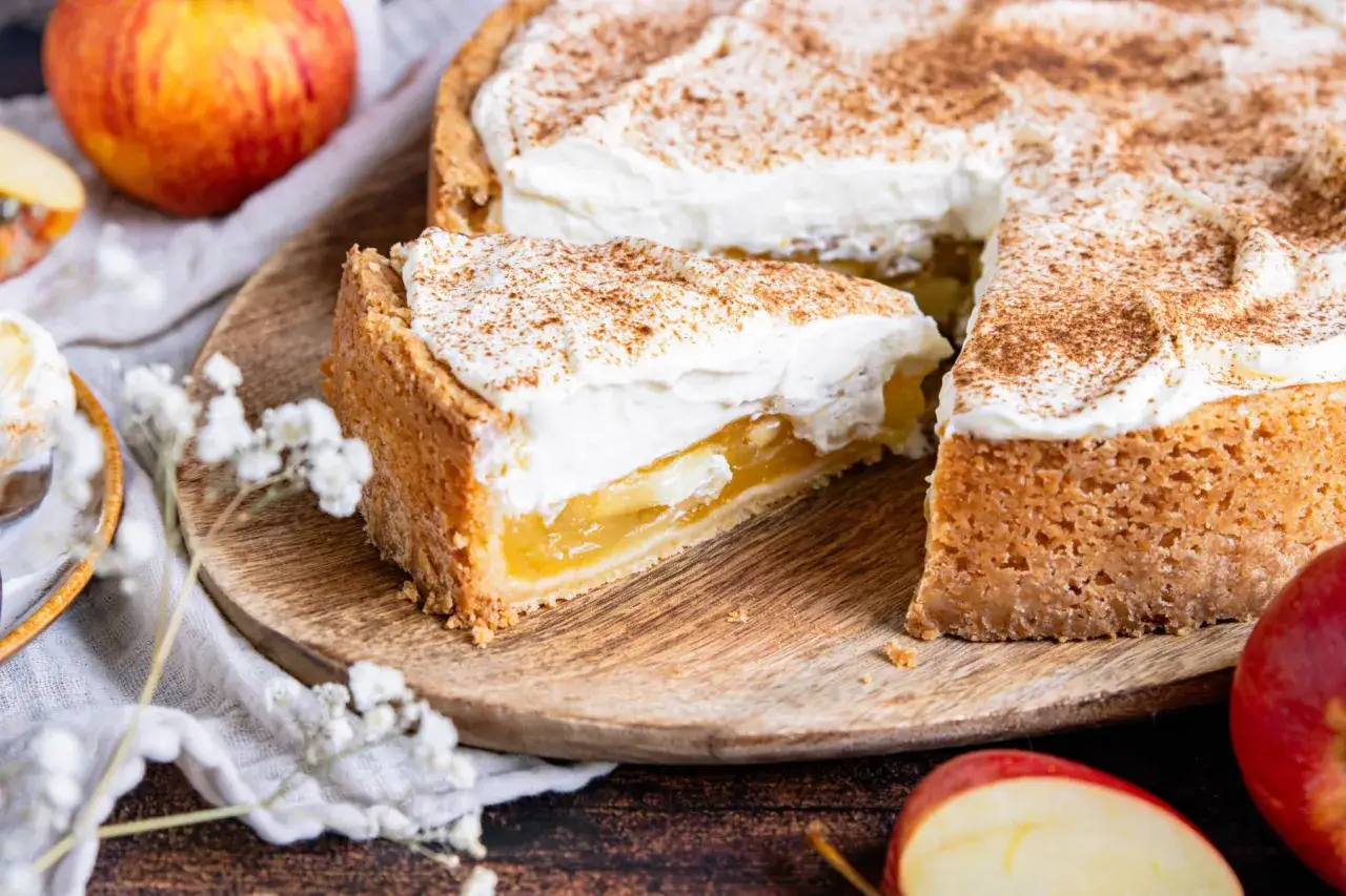 Ein St&uuml;ck Apfel Schmand Torte mit cremigem Topping und Zimt bestreut, daneben &Auml;pfel und zarte Bl&uuml;ten.
