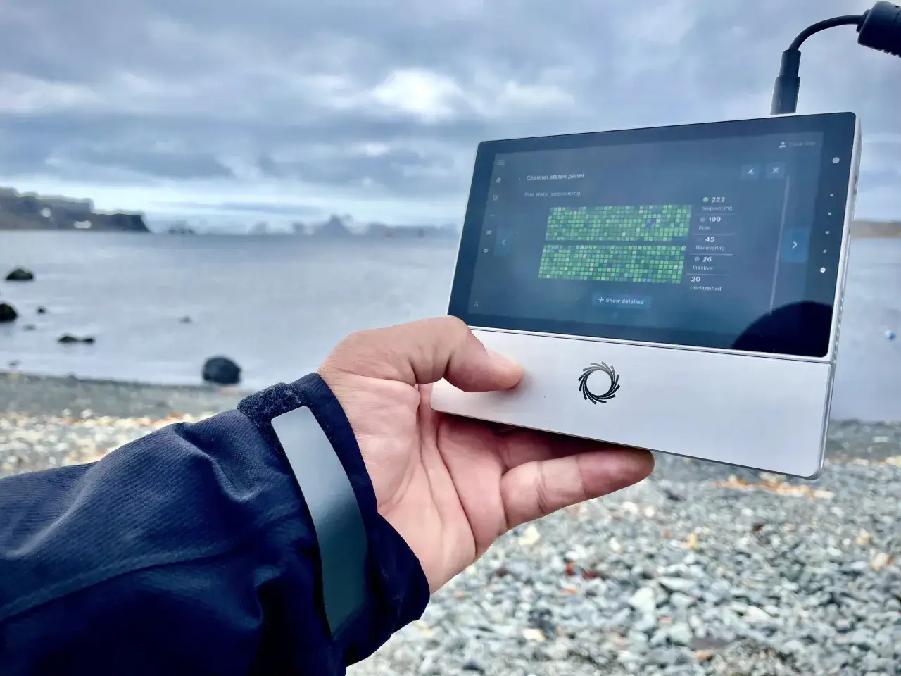 Mano sostiene un dispositivo MinION Nanopore en la playa, mostrando datos de secuenciaci&oacute;n.