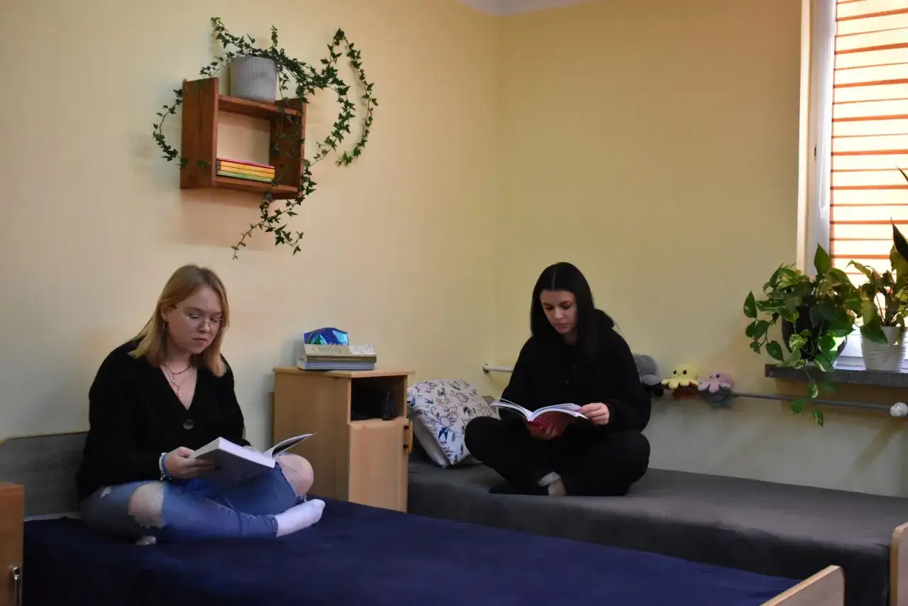 Dwie studentki w pokoju, jedna czyta książkę, druga przygotowuje się pedagogicznie do zajęć.